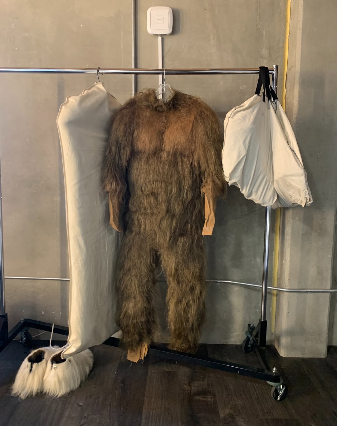 Furry brown costume hangs on a metal rack between two garment bags in front of a gray wall. The costume has fur-like material.